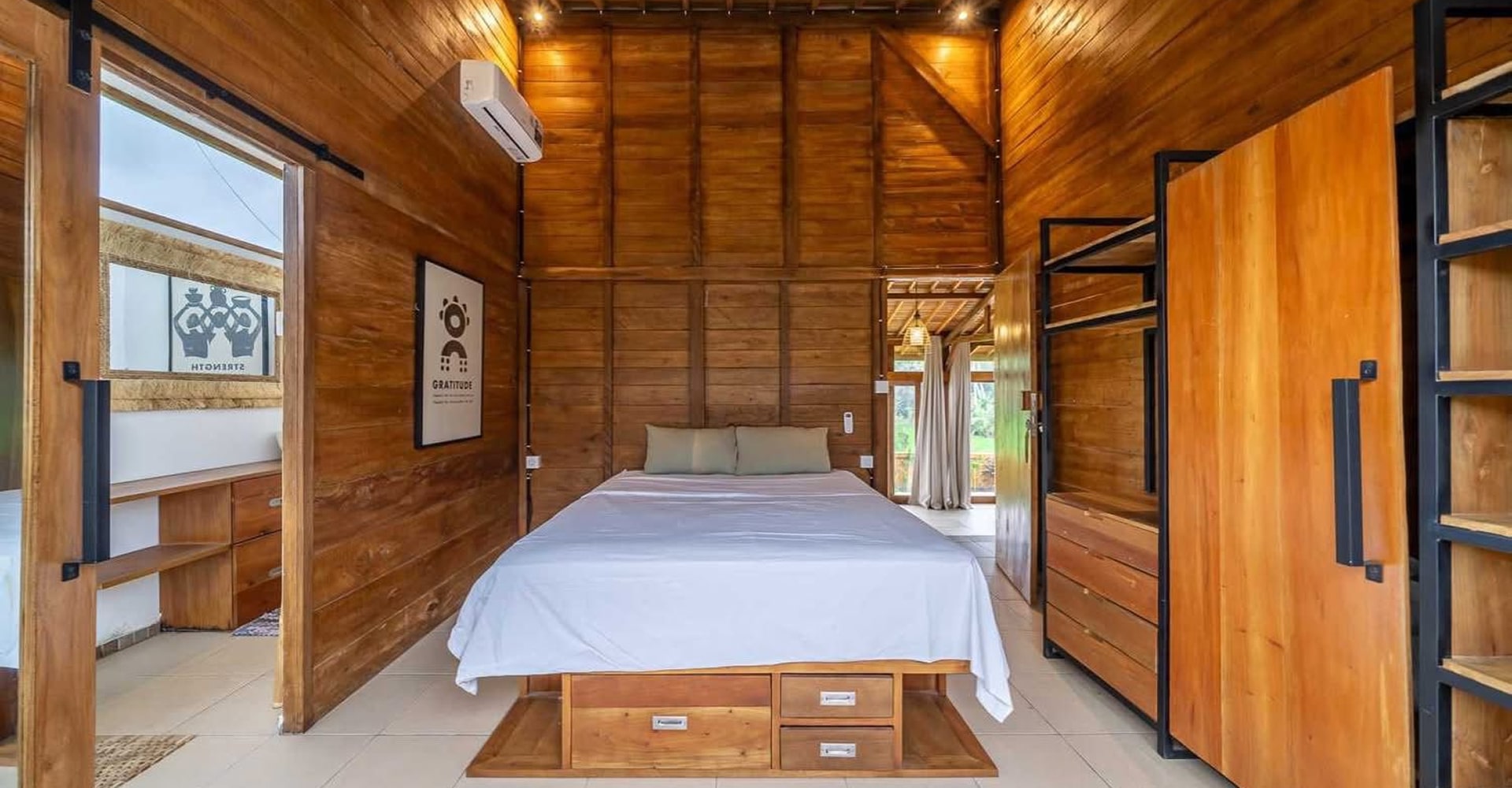 Chambre cocooning en bois avec grand lit blanc et éclairage chaleureux; affiches "GRATITUDE" et "STRENGTH".