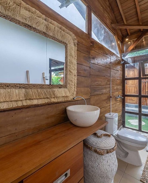 Salle de bain rustique avec murs et plafond en bois, miroir à franges en paille et vasque ronde en pierre au-dessus d'un meuble bois.